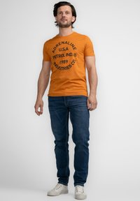 Camiseta de algodón naranja con texto gráfico negro que dice "ADRENALINE U.S.A PETROL IND. 1989 INDUSTRIES CO." Combinada con jeans de mezclilla azules y zapatillas blancas.
