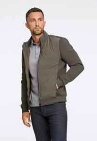 Olijfgroen jack met een doorgestikte voorkant, ritssluiting en geribde mouwen. Draag het over een grijze poloshirt en gecombineerd met donkere jeans.
