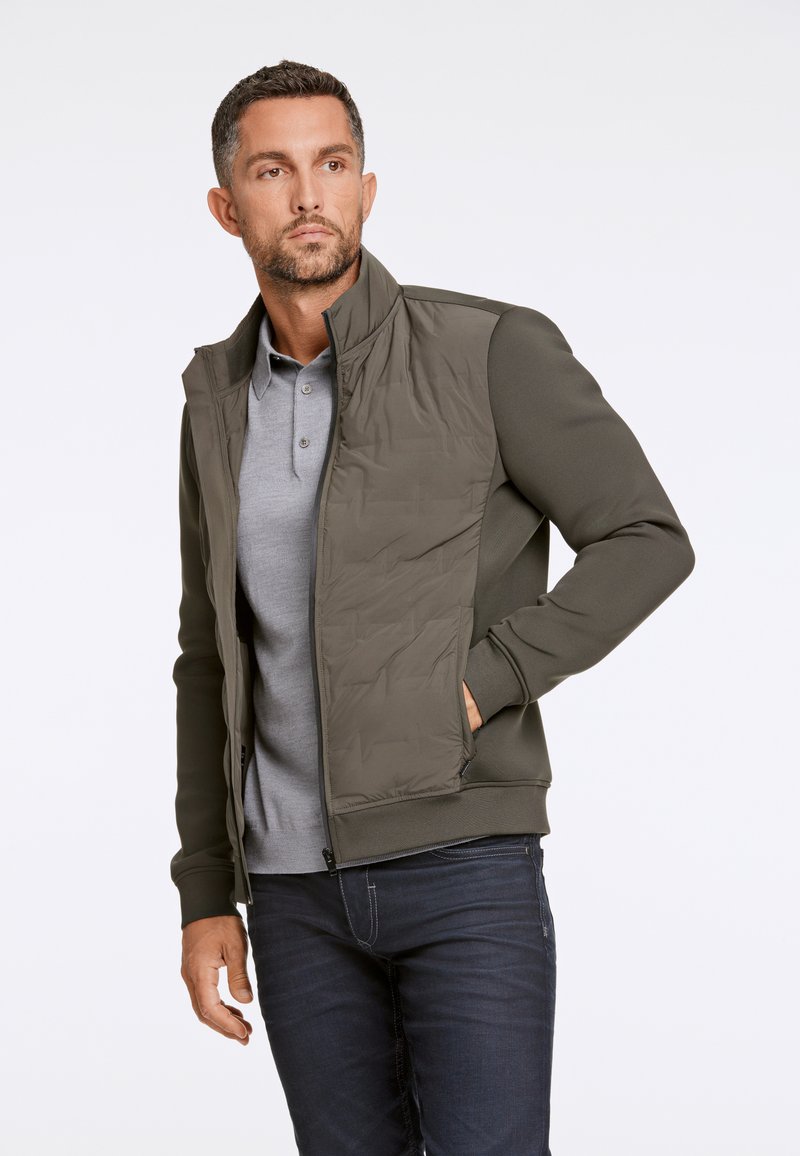 Olijfgroen jack met een doorgestikte voorkant, ritssluiting en geribde mouwen. Draag het over een grijze poloshirt en gecombineerd met donkere jeans.