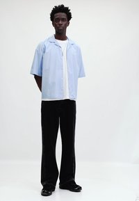 Camicia azzurra chiara a maniche corte sopra una maglietta bianca, abbinata a pantaloni neri a gamba larga e scarpe nere lucide, in piedi su uno sfondo neutro.