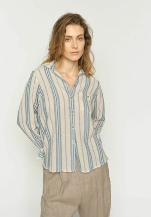 Mlada ženska z valovitimi lasmi, oblečena v bež in modro črtasto srajco z gumbi ter široke bež hlače, stoji pred enostavnim ozadjem.