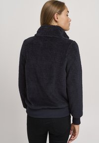 Marinefarget fleecejakke med høy krage, teksturert stoff, ribbede mansjetter og hem, og ingen synlige lukkinger. Enkel, uformell design.