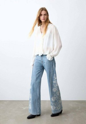 Mujer que lleva una blusa blanca translúcida y vaqueros de pierna ancha azul claro con bordados florales blancos, de pie sobre un suelo gris contra una pared blanca.