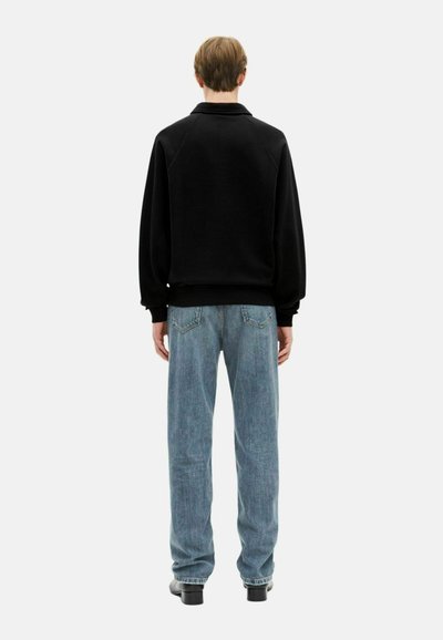 Asmuo stovi nugara į mus, apsirengęs juodu megztiniu, šviesiai mėlynomis tiesiomis kelnėmis ir juodais batais, priešais paprastą baltą fono.