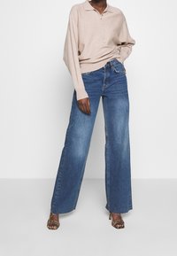 Top lavorato a maglia color beige chiaro con colletto e patta con bottoni, abbinato a jeans a gamba larga di colore denim medio. L'outfit è completato da sandali con cinturini.