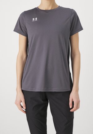 Henkilö, joka pukee lyhythihainen, tummanharmaa Under Armour -urheilupaita ja mustat housut, seisoo yksinkertaisen taustan edessä.