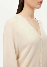 Beige kofta med V-ringning, prydd med en rad ljusa knappar och fin ribbad textur längs framknäppningen och ärmarna.