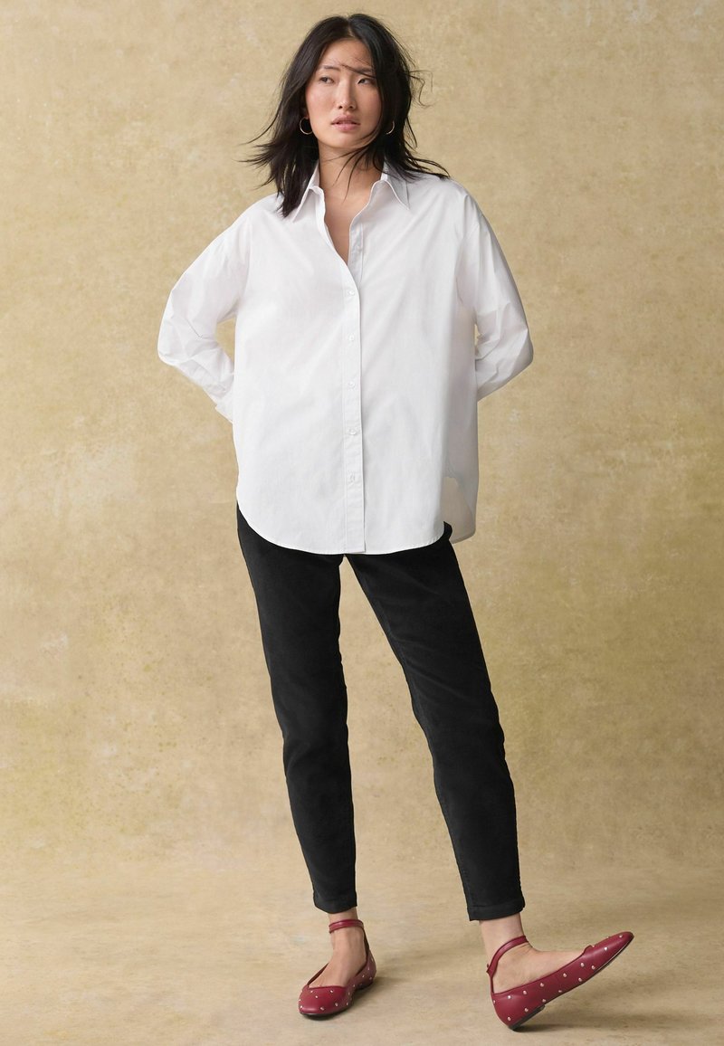 Camicia bianca con bottoni, pantaloni neri slim fit e balerine rosse a pois bianchi. La camicia ha una vestibilità comoda, un orlo arrotondato e maniche lunghe.