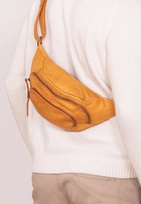 Geltonas odinis fanny pack'as su kreiviu, dviem užtrauktukais uždaromais kišenėmis, reguliuojamu diržu ir lygiu paviršiumi, nešiojamas per kūną.