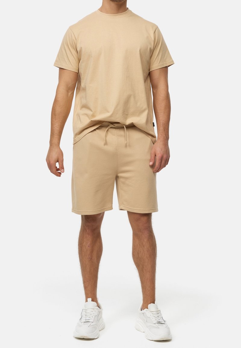 Homme portant un T-shirt beige à manches courtes et un short assorti à cordon, associé à des baskets blanches, debout devant un fond uni.