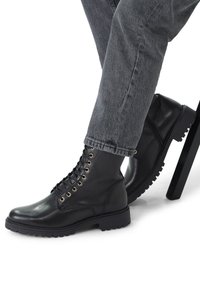 Des bottes noires en cuir à lacets présentent des œillets dorés, une semelle crantée et des détails de couture. Associées à un jean gris retroussé.