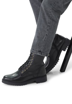 Des bottes noires en cuir à lacets présentent des œillets dorés, une semelle crantée et des détails de couture. Associées à un jean gris retroussé.