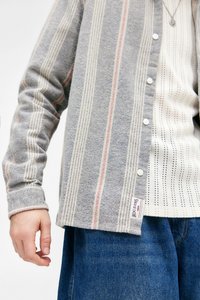 Gråstripet skjorte med knapper, med røde og hvite linjer, sammen med en hvit teksturert undertrøye. Har en logomerke på kanten. Blå jeans synlige.