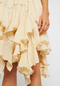 Light beige dress with layered, ruffled design. Silver trim accents along the edges. Soft, textured fabric with a flowing appearance.