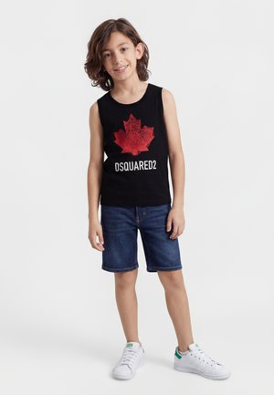 Smilende dreng med skulderlangt hår iført sort ærmeløs trøje med rød ahornblad og "DSQUARED2", cowboyshorts og hvide sneakers.