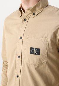 Béžová košile na zapínání vyrobená z bavlny. Má náprsní kapsu s černou štítkem zobrazujícím "CK" a "Calvin Klein Jeans." Jednoduchý design.