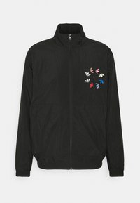 Svart vindjacka med dragkedja framtill, med flerfärgade broderade lövmönster på övre vänstra sidan och elastiska ärmslut.