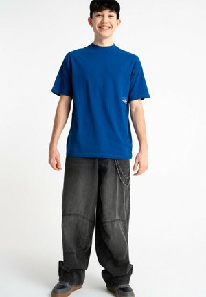 Młody mężczyzna uśmiechający się, ubrany w niebieską koszulkę, czapkę z daszkiem, luźne ciemne jeansy z łańcuchem oraz szare trampki, stojący na białym tle.