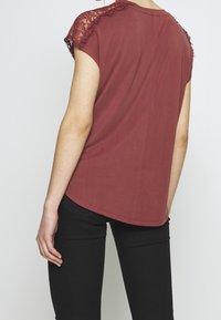 Burgundské tričko s krajkovými detaily na ramenou, krátkými rukávy, zaobleným lemem a měkkou texturou; kombinované s černými upnutými kalhotami.