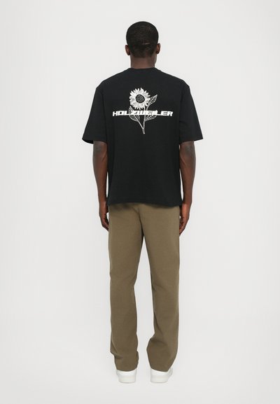 Schwarzes Baumwoll-T-Shirt mit einem weißen Sonnenblumen-Graphic und Text auf dem Rücken, kombiniert mit olivgrünen Hosen und weißen Sneakers.