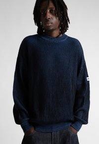 Jaunas vyras su dreadsais, apsirengęs juostuotu tamsiai mėlynu ir juodu megztiniu bei tamsiomis džinsomis, rankos kišenėse, stovi prieš vienodą fono.