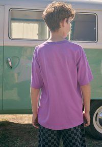 Paarse katoenen T-shirt met korte mouwen, ronde zoom, gedragen met zwart en grijs geblokte broeken. Achtergrond toont een groene bus.