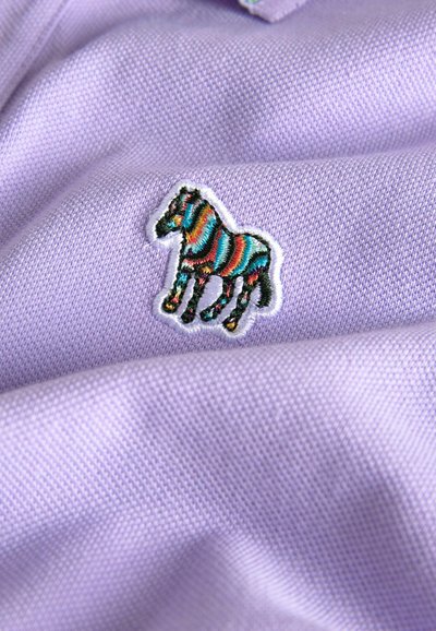 Lavendel polo skjorta med en färgglad broderad zebra patch med vit kontur, mot en texturerad tygbakgrund.