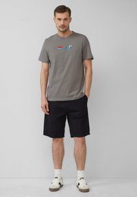 Szary bawełniany t-shirt z grafiką "JAWS" i wzorem fal, w połączeniu z czarnymi szortami do kolan i białymi niskimi trampkami.