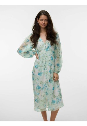 Žena s dlhými tmavými vlasmi nosí priesvitné, dlhorukávové midi šaty s kvetinovým vzorom v modrej a zelenej farbe na bielom pozadí.