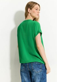 Blusa verde testurizzata a maniche corte con una vestibilità rilassata, abbinata a pantaloni corti in denim blu. Il tessuto presenta un sottile motivo ondulato e una leggera lucentezza.