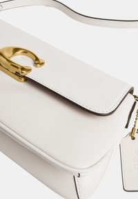 Sac à bandoulière en cuir blanc avec une fermeture en métal doré et une bandoulière blanche cousue assortie, avec une étiquette de marque sur une chaîne à billes.