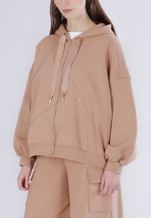 Donna che indossa una felpa beige oversize con zip e pantaloni coordinati con piccoli ornamenti a forma di stelle e punti.