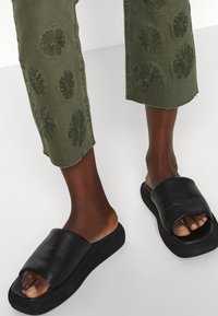 Chinelas pretas com uma tiras largas na parte superior, apresentando uma textura mate. Calças verde-azeitona com bordados florais e bordas desfiadas na bainha.