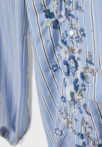 Modrá a biela pruhovaná látka s detailnou modrou, bielou a striebornou flitrovou výšivkou kvetov na dlhom rukáve.