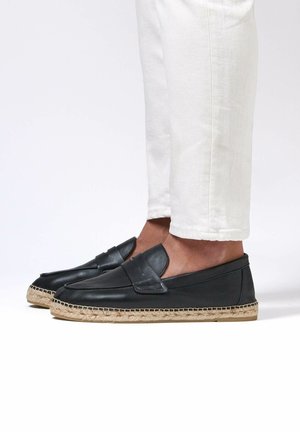 Person trägt schwarze Leder-Slipper mit gewebten Jute-Sohlen und weißen Hosen, die oberhalb des Knöchels gekürzt sind, und steht auf einer weißen Fläche.