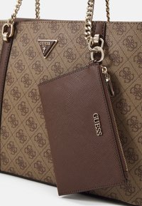 Sac fourre-tout Guess marron avec motif monogramme, anses en chaîne dorée, et poche zippée texturée marron attachée avec le logo Guess.