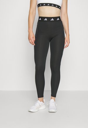 Czarne legginsy o wysokim stanie i teksturowanej tkaninie, z białą taśmą z logo na górze oraz bocznymi paskami dla uzyskania detalu. Noszone z białymi sneakersami.