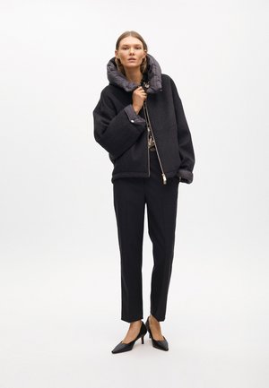 Chaqueta negra de mezcla de lana con capucha acolchada gris, cremallera plateada y dos bolsillos laterales, combinada con pantalones negros a medida y tacones.