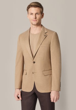 Blondyn w beżowej marynarce na beżowej koszuli i ciemnobrązowych spodniach, stoi na tle jednolitego jasnobeżowego tła.