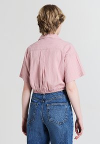Camicia a maniche corte color rosa chiaro con righe verticali, dettagli di pieghe sul retro e orlo arricciato, abbinata a jeans blu a vita alta.