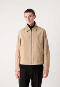 Beige leather jacket with a zip front, stand collar, and two side pockets, paired with a black ribbed turtleneck underneath.