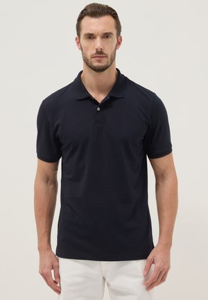 Hombre con una camisa polo negra de manga corta y pantalones blancos, de pie frente a un fondo claro y liso.