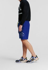 Pantalones cortos azules con texto blanco impreso, combinados con sudadera negra con letras blancas. Los zapatos son multicolores con suelas texturizadas.