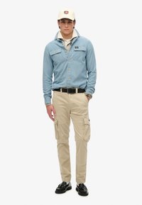 Lichtblauwe denim overhemd met voorzakken, beige cargobroek met zijzakken, en zwarte instappers. Beige pet met logo.