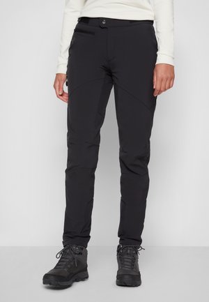 Personne portant un pantalon de plein air noir et des chaussures de randonnée noires, debout devant un fond uni clair.