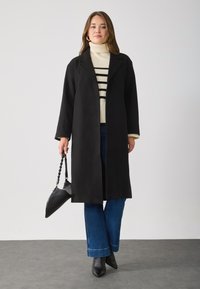Cappotto lungo nero in lana con collo a risvolto; abbinato a un dolcevita a righe crema, jeans blu a zampa d'elefante e stivaletti neri con punta.
