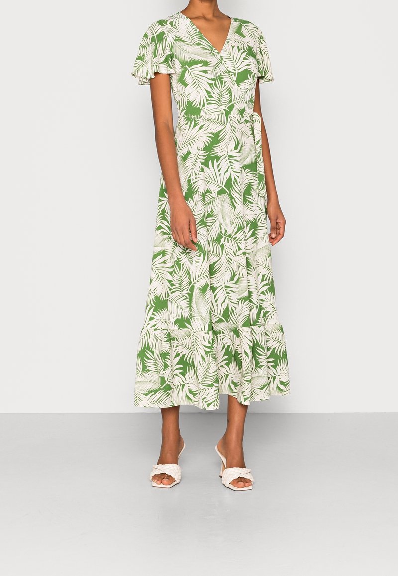 Woman wearing a green midi wrap dress with white tropical leaf print and short flutter sleeves, paired with white heeled sandals.