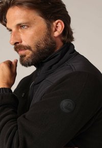 Zwarte hybride jas met een gladde bovenkant en een zachte fleece onderkant. Ritsluiting en een ronde logo patch op de linker mouw.