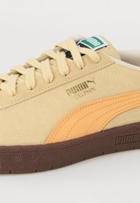 Baskets en daim beige avec une semelle texturée marron, une bande orange sur le côté et une étiquette logo verte. Fermeture à lacets avec lacets plats beige.