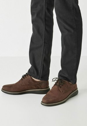 Brune ruskinds snøresko med rund toe, tekstureret overflade og en kontrasterende sort gummisål, parret med mørke jeans.